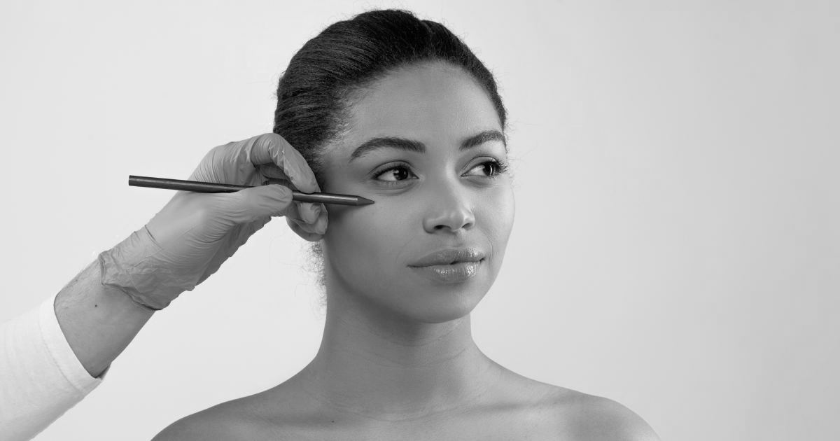 Aesthetic medicine, injection cosmetology concept. Plastic surgeon hands in medical gloves holding pen, making preparation lines on beautiful young black woman face, isolated on grey background